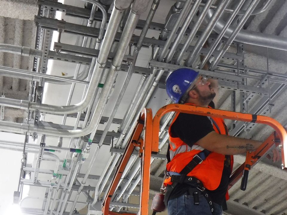 Técnico sobre plataforma de elevación revisando canalizaciones y soportes de cableado en el techo de un cuarto eléctrico.