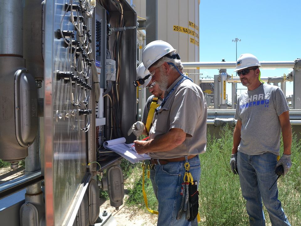Dos técnicos revisan el cableado de un tablero de control en el exterior de una planta industrial.